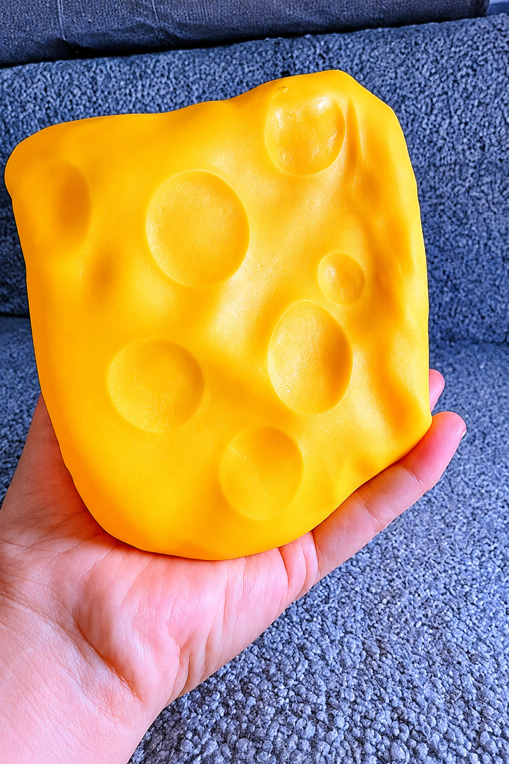 A hand holding a yellow, cheese-like squishy toy with embossed circular holes, photographed against a dark textured background.