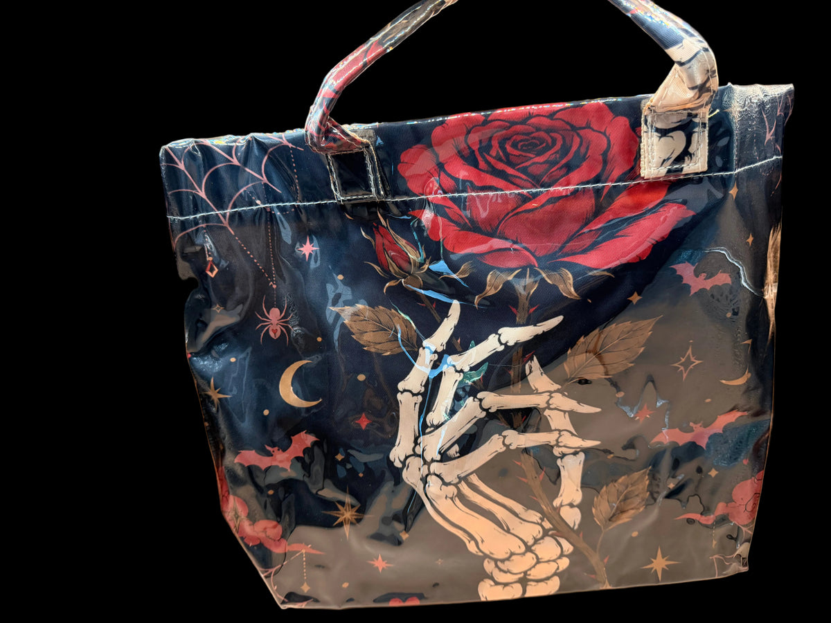 A glossy black tote bag featuring a gothic skeleton hand graphic reaching up toward a large red rose, with stars, moons, bats, and spiders printed across the surface. The bag has silver-toned metal hardware and is photographed against a reflective metallic background.